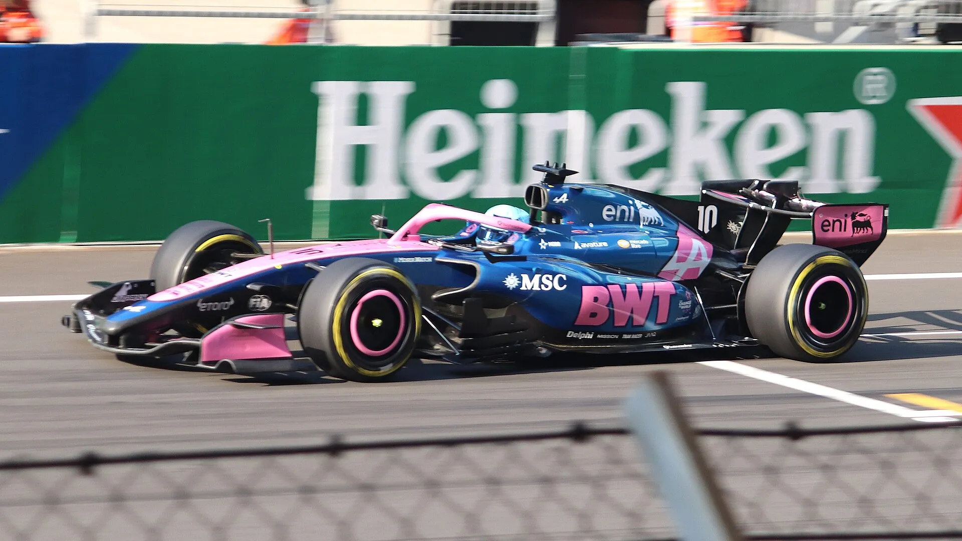 pierre gasly's pink and blue alpine at speed in shanghai, seen over a catch fence