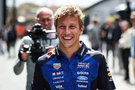 liam lawson laughing in a red bull team shirt in the paddock, a cameraman behind him