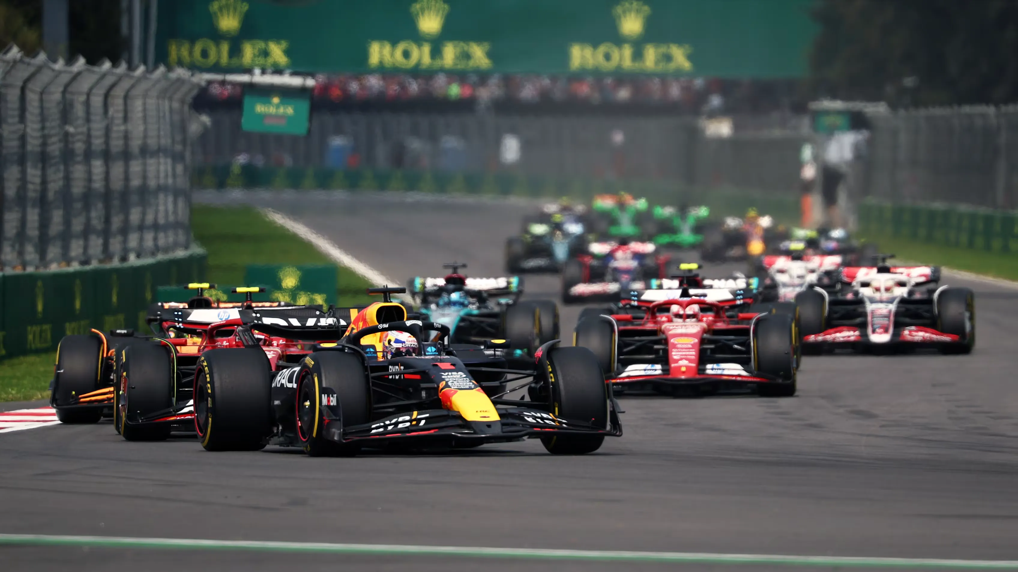 a red bull and two ferraris run side by side towards turn 1 in mexico city