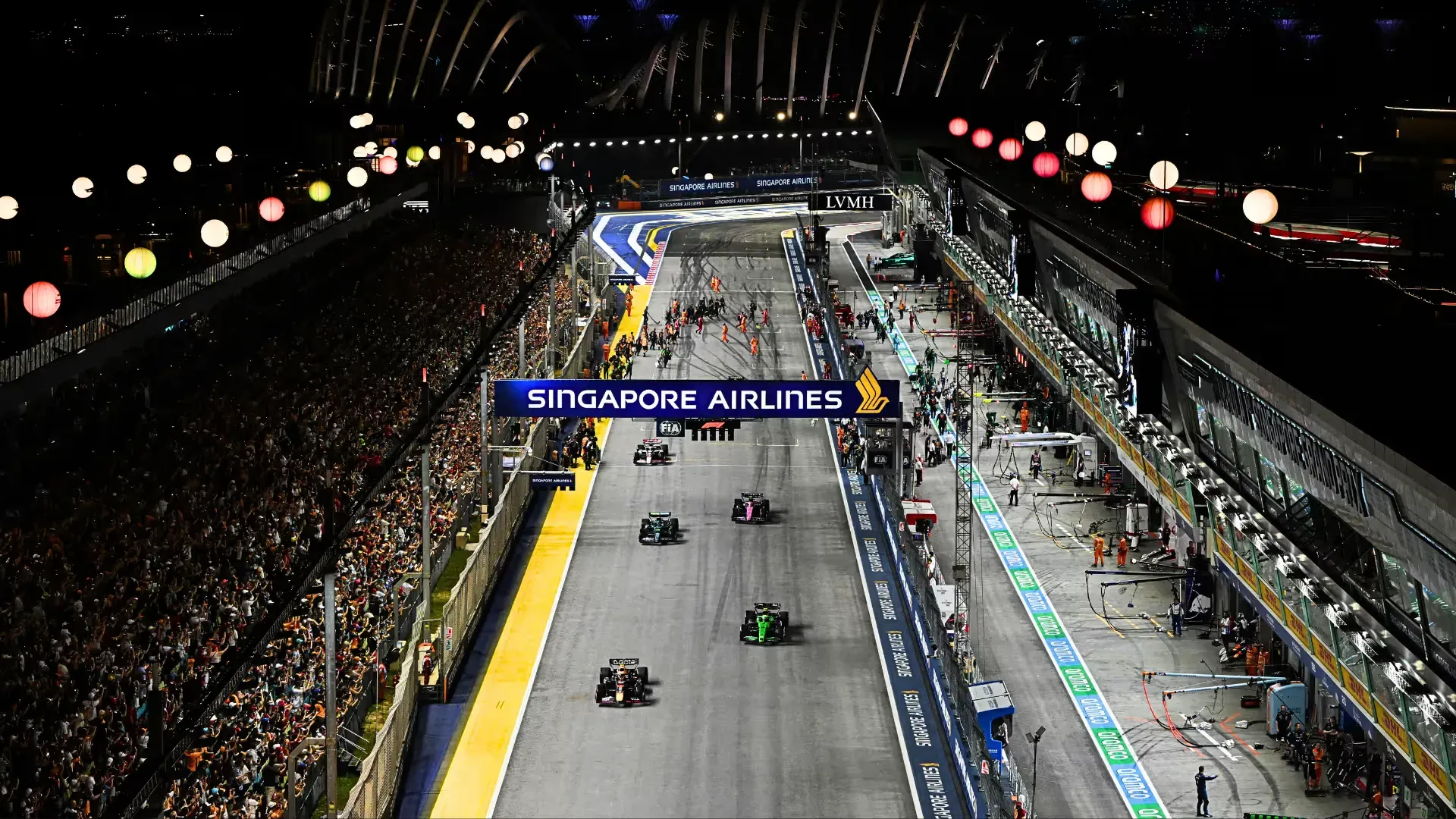 a red bull leads a line of cars down the floodlit pit straight at marina bay, past packed grandstands